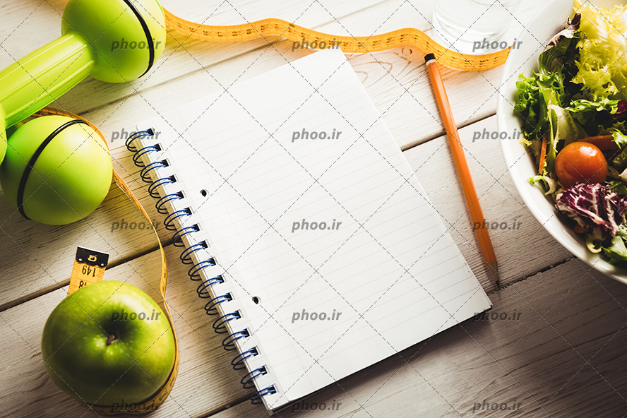 عکس با کیفیت متر و دمبل و سیب در کنار دفترچه و مداد و ظرف سالاد در گوشه تصویر روی میز چوبی