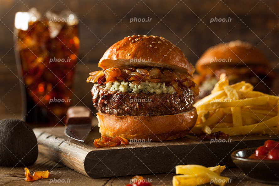 عکس با کیفیت ساندویچ همبرگر در کنار لیوان نوشابه و سیب زمینی بر روی تخته چوبی