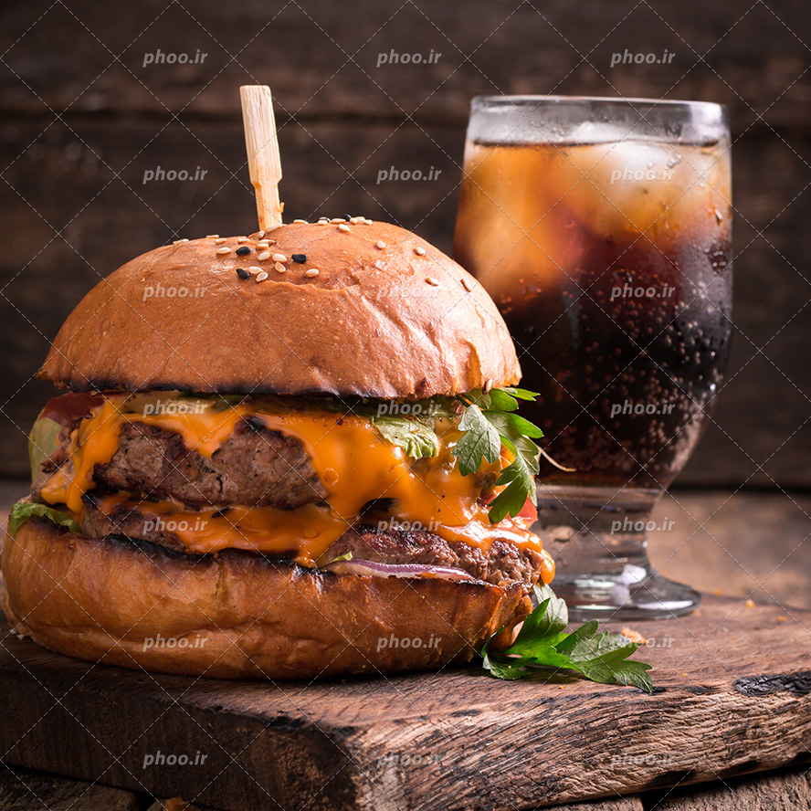 عکس با کیفیت ساندویچ همبرگر در کنار لیوان نوشابه یخ از نمای نزدیک قرار گرفته بر روی تخته چوب