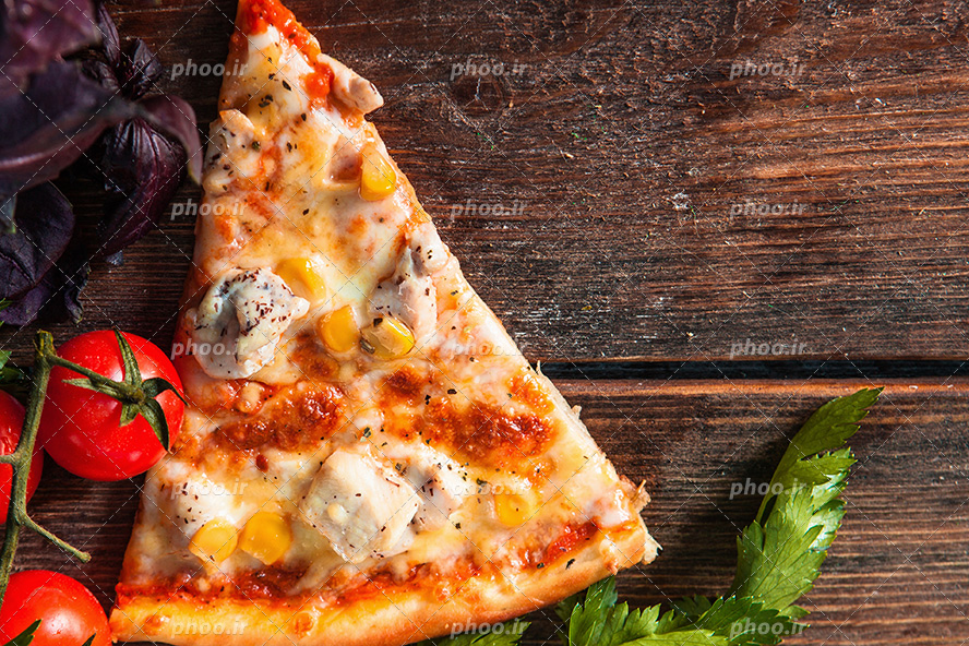 عکس با کیفیت یک اسلایس پیتزا در کنار گوجه و سبزیجات قرار گرفته بر روی میز چوبی