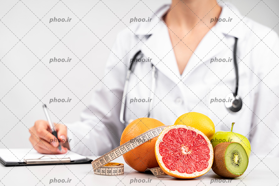 عکس با کیفیت پزشک متخصص تغذیه با روپوش سفید در حال نوشتن برنامه ی غذایی و میوه های تازه و مفید بر روی میز