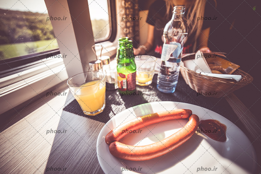 عکس میز صبحانه کنار پنجره که روی پنجره صبحانه شامل آب پرتغال و سوسیس است