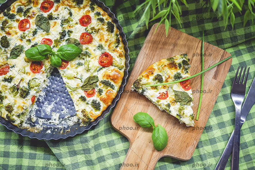 عکس پیتزا سبزیجات در سینی فر روی میز آشپزخانه با تخته چوبی که روی آن یک قاچ پیتزا قرار داده شده است