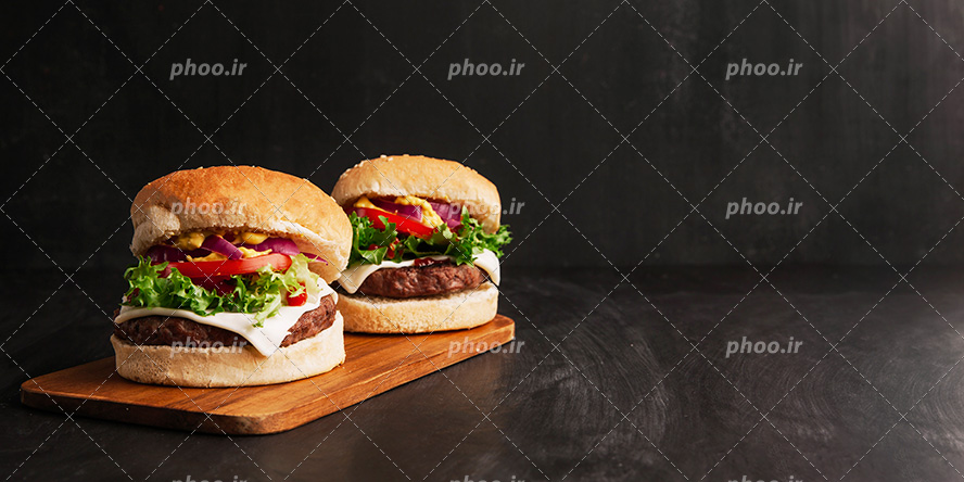 عکس با کیفیت دو همبرگر خوشمزه در کنار یکدیگر بر روی تخته چوب و پس زمینه به رنگ مشکی