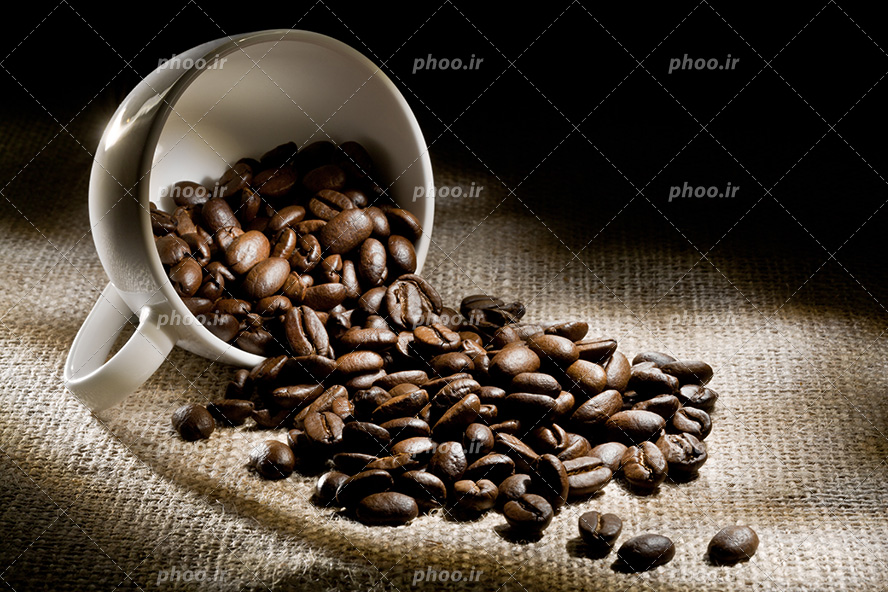 عکس با کیفیت دانه های ریخته شده قهوه بر روی پارچه ی کنفی از داخل فنجان سفید