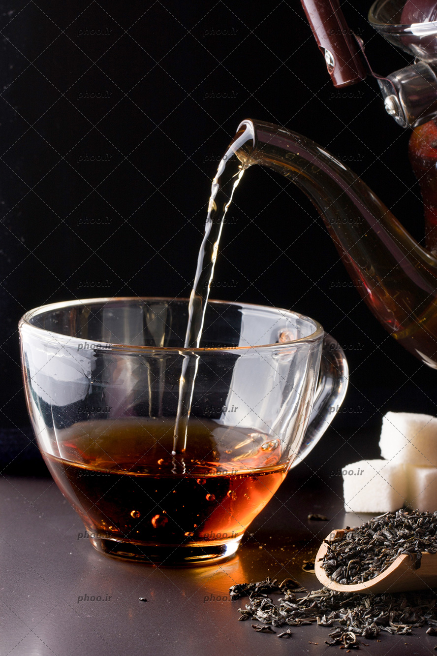 عکس ریختن چای از قوری به فنجان در کنار قاشق چوبی با چای خشک و قند در پس زمینه مشکی