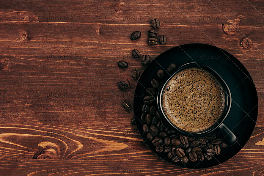 عکس فنجان قهوه روی نعلبکی در کنار دانه های قهوه در پس زمینه چوبی