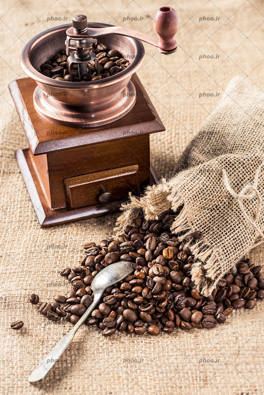 عکس آسیاب قدیمی قهوه در کنار دانه های قهوه روی زمین و داخل گونی در پس زمینه پارچه کنفی