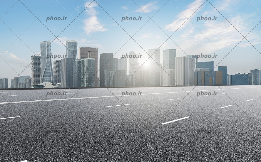 عکس بزرگراه در کنار شهر با ساختمان های بلند در برابر آسمان آبی و خورشید