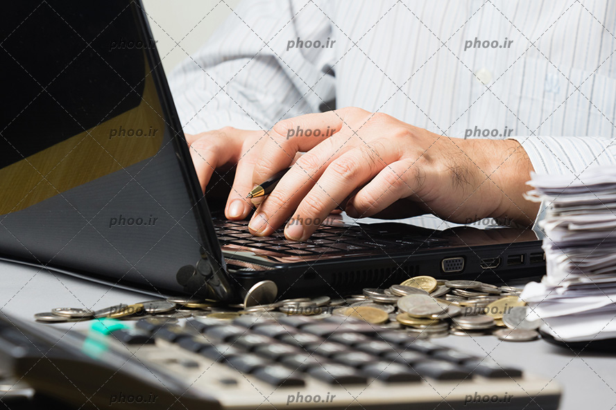 عکس دستان در حال تایپ در لپ تاپ و سکه ها و کاغذ ها